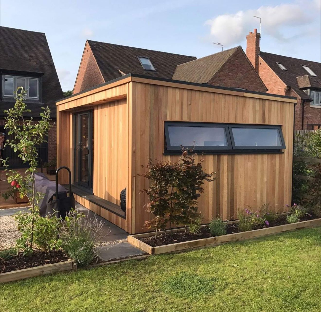 Modern wooden garden office with dark windows and door, set in a yard with a brick house backdrop.