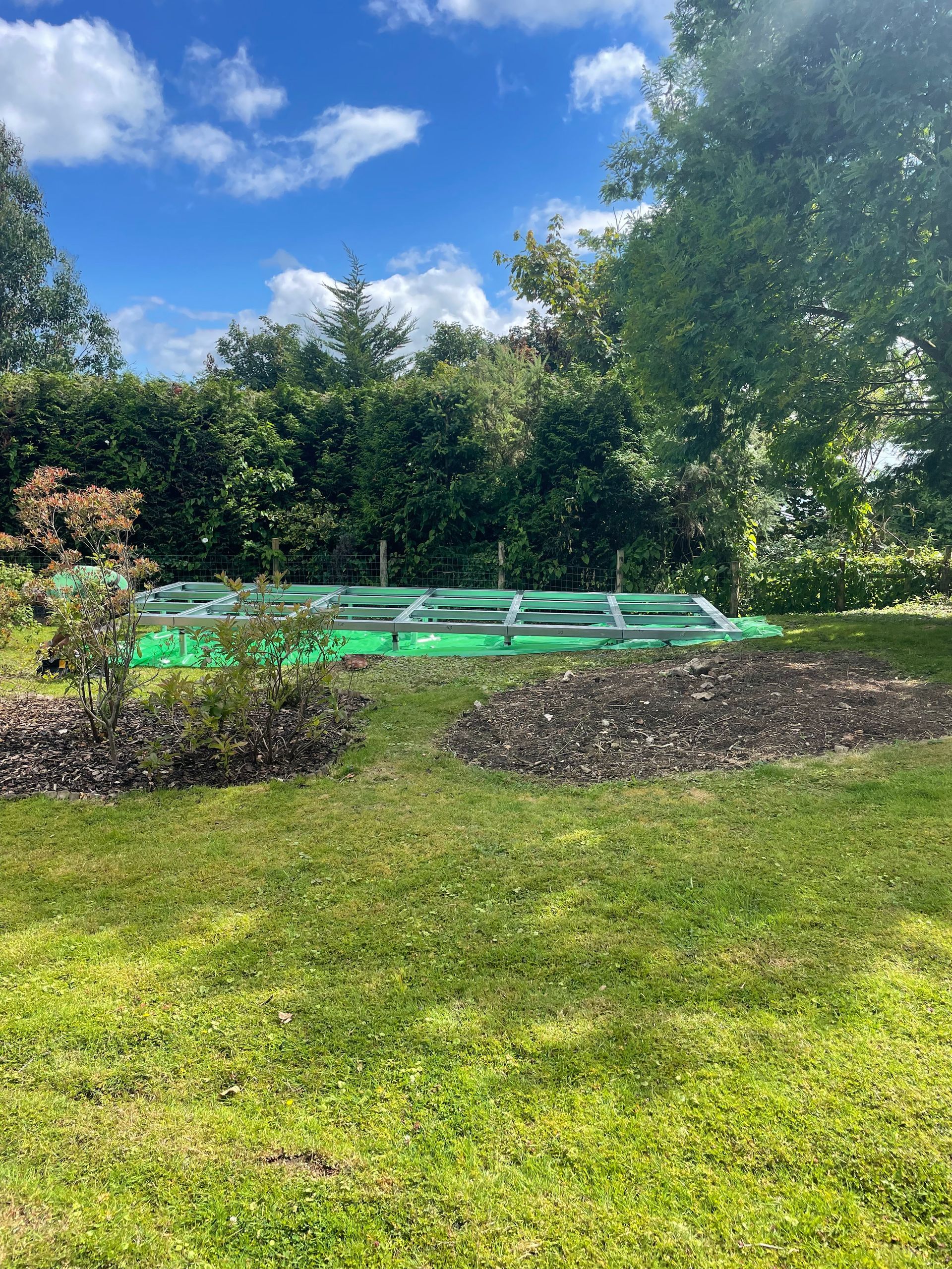 Green garden beds with metal frames sit on green grass, under a blue sky with trees in the background.