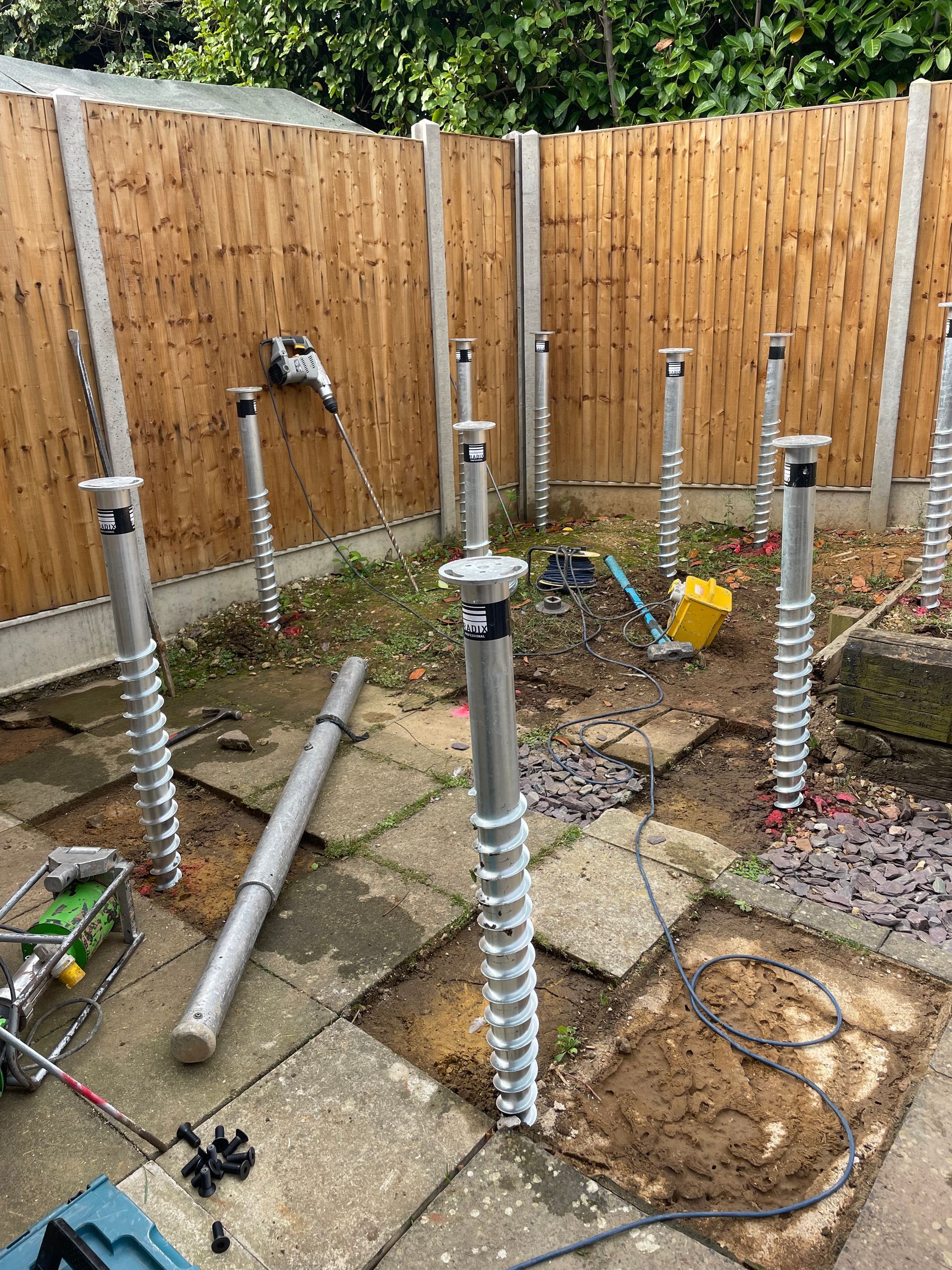 Metal ground screws installed in a backyard, ready for construction. A drill is visible.