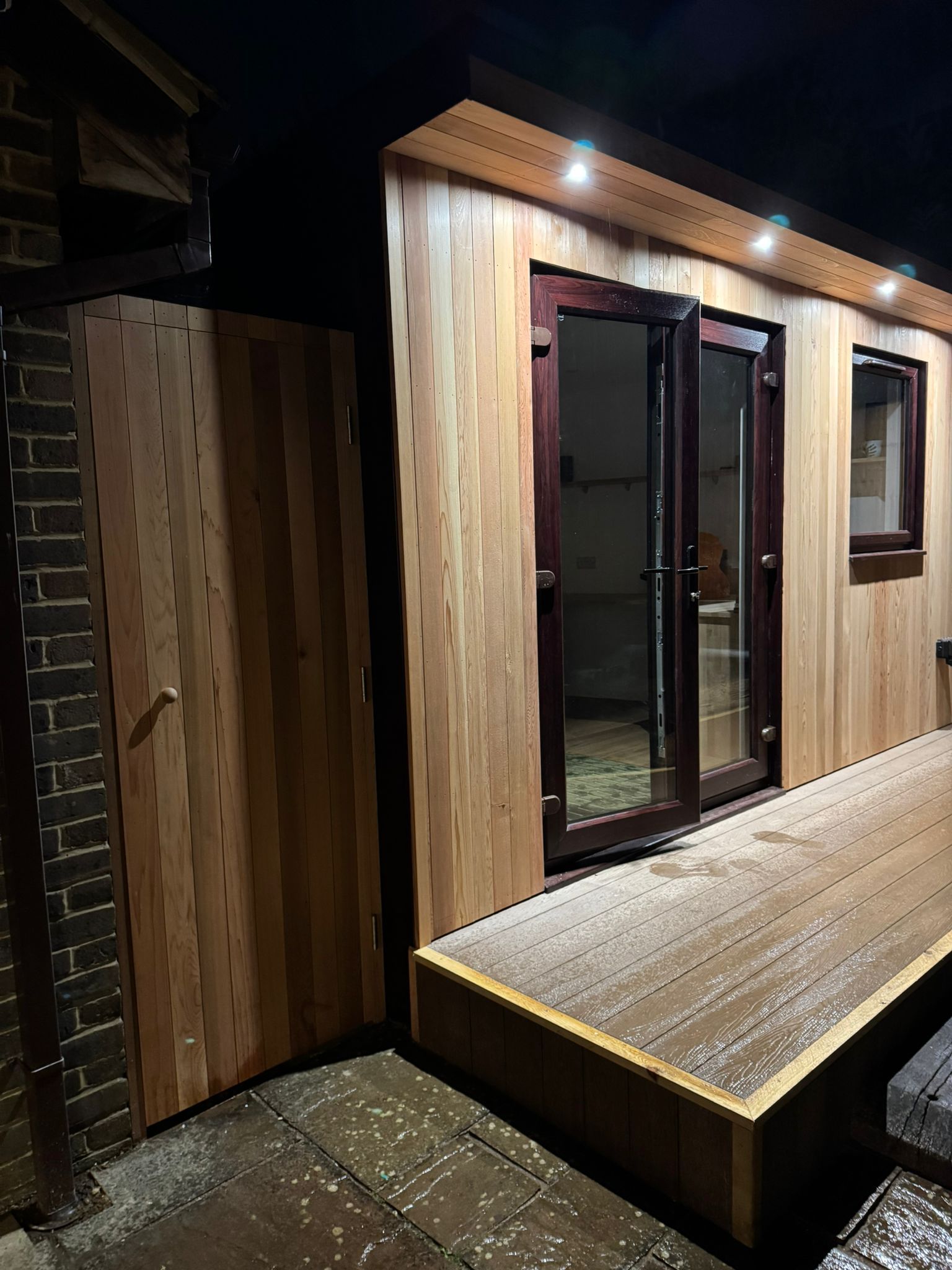 Wooden garden office with double doors, small window, and a gate at night.