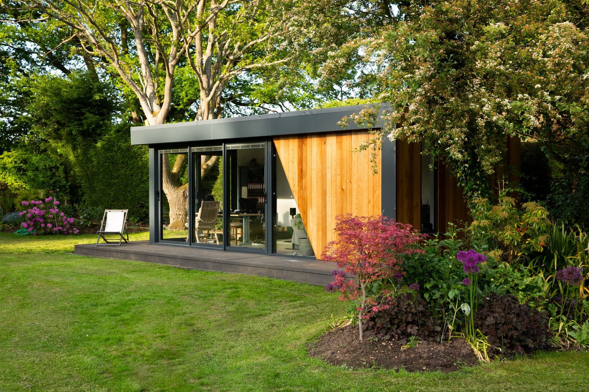 Modern backyard office building with glass doors and wood siding, surrounded by lush green lawn and garden.