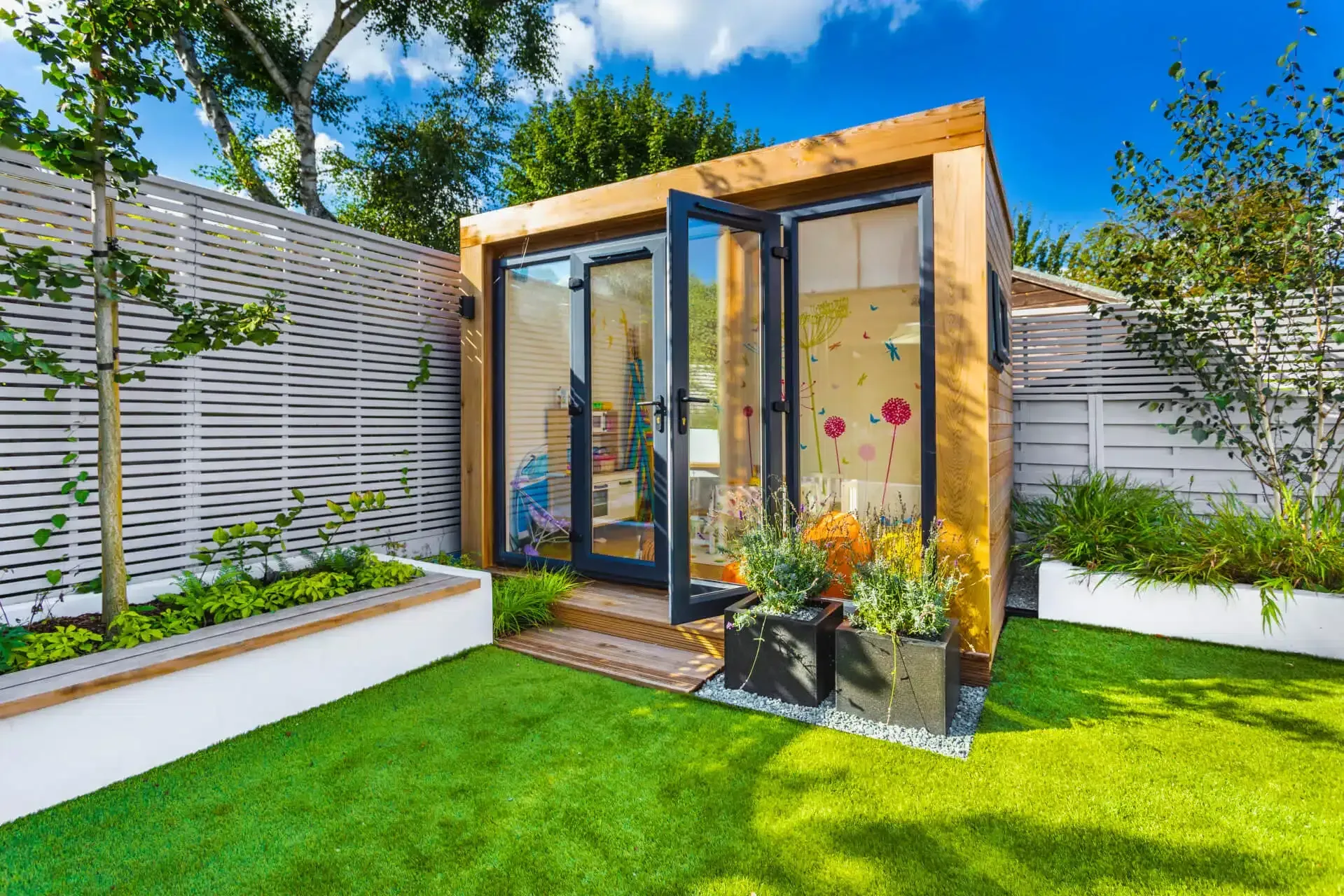 Wooden garden office with glass doors, surrounded by greenery and white fence under a blue sky.