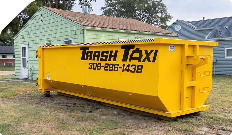 Yellow Trash Taxi dumpster in a yard beside a house, with phone number printed on the side.