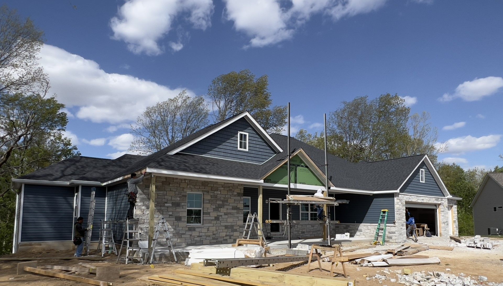 House under construction with gray siding, exposed framing, and workers' scaffolding under a partly cloudy sky