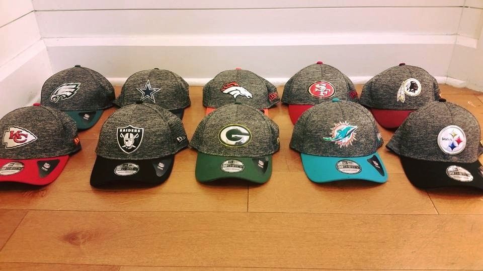 Ten NFL team hats with speckled grey crowns arranged in two rows on a wooden floor against a white wall.
