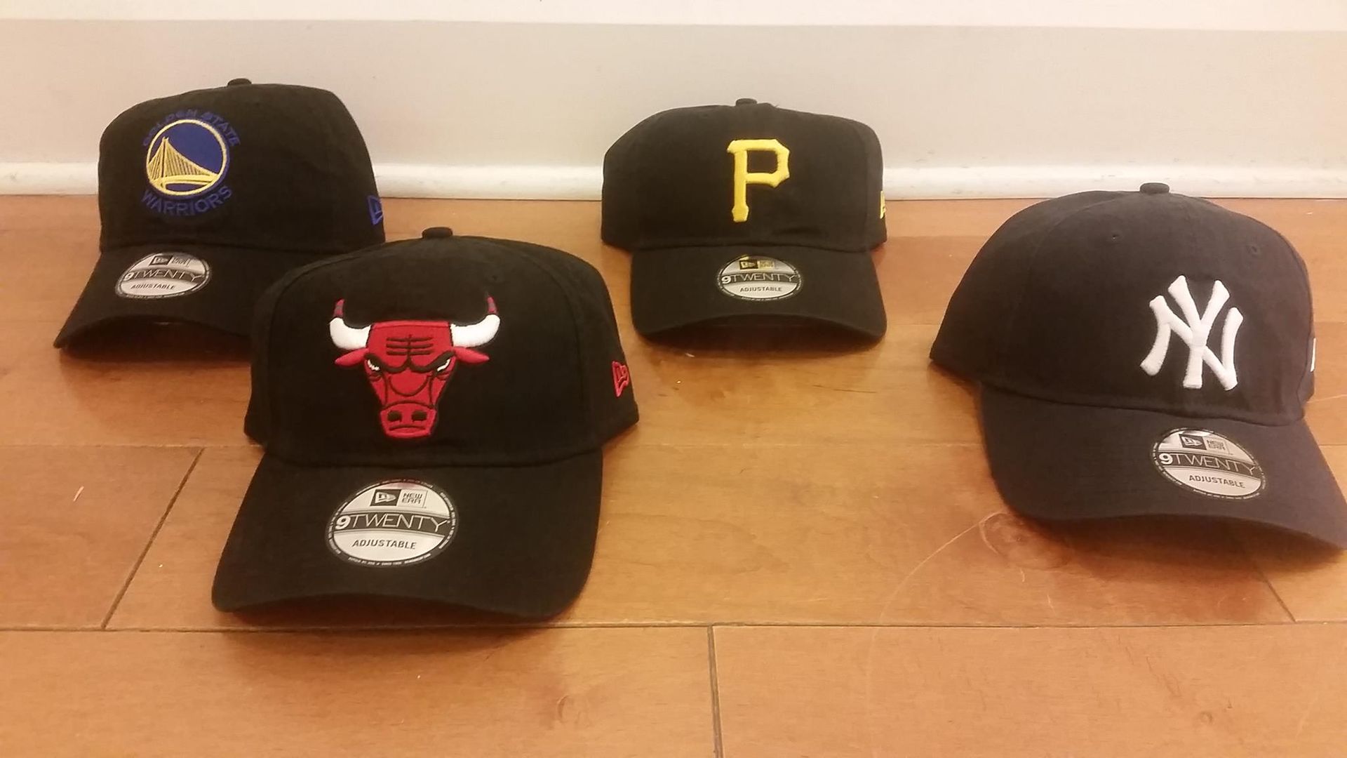 Four black sports team baseball caps featuring the Warriors, Bulls, Pirates, and Yankees logos on a wood floor.