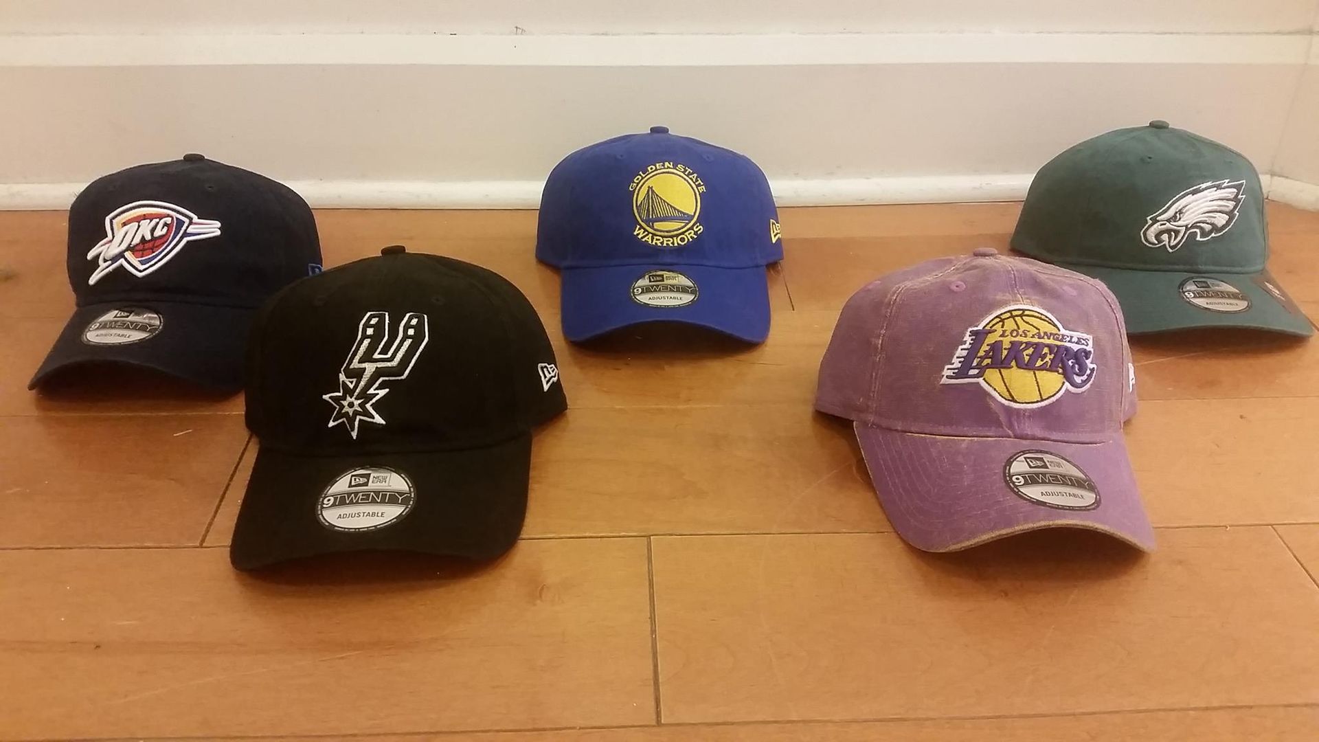 Five sports team baseball caps arranged on a wooden floor, featuring various team logos and colors.