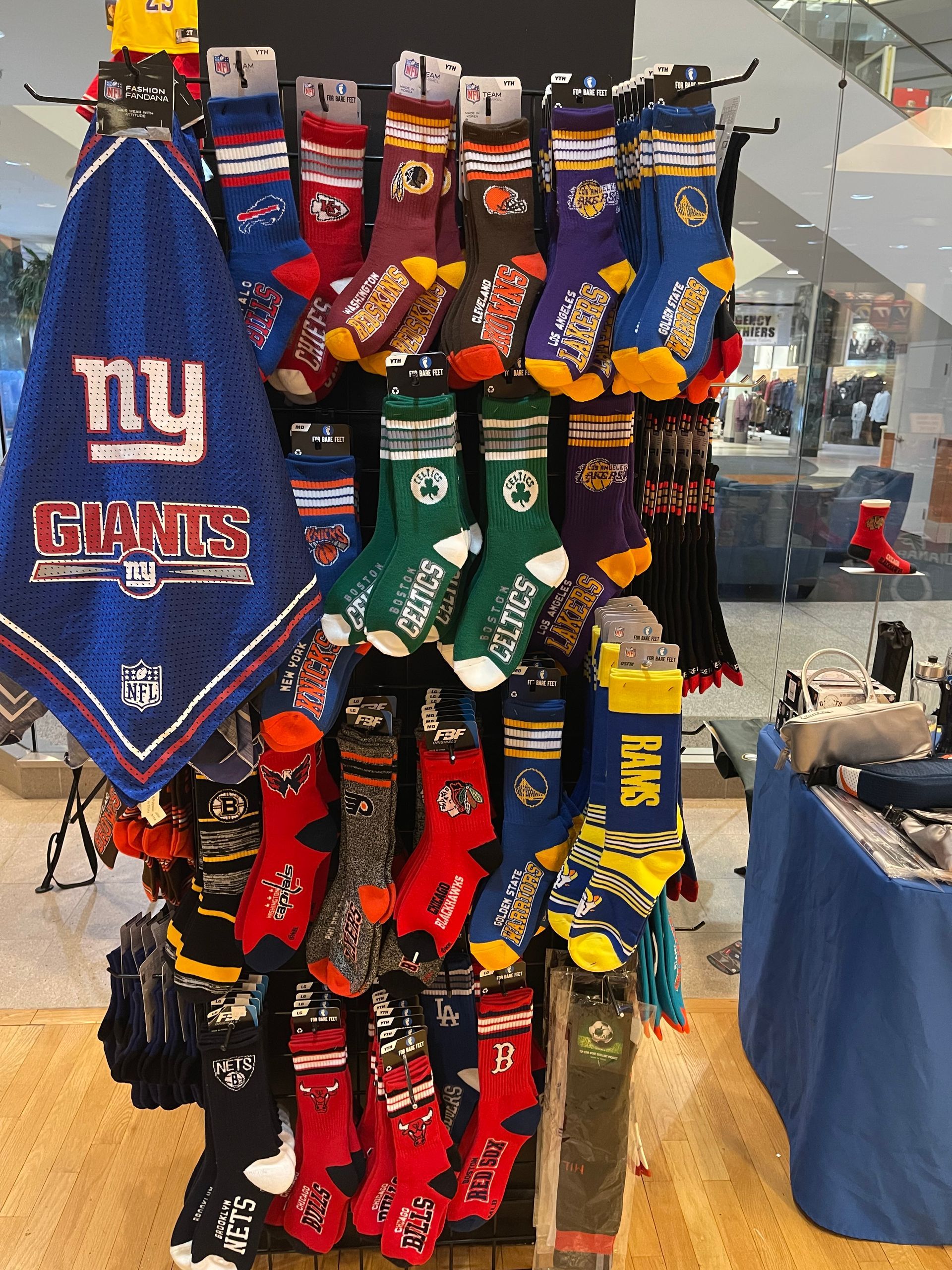 A display rack holding various sports-themed socks and a blue New York Giants bandana in a retail store setting.