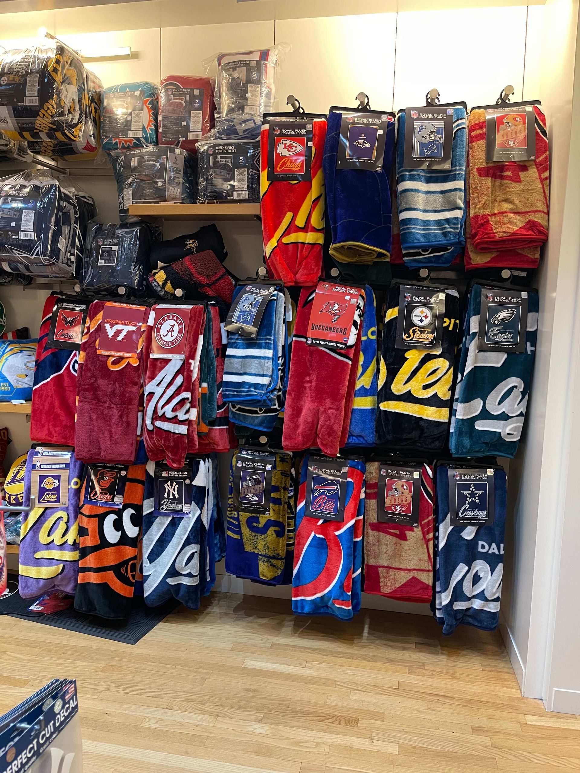 A retail display of folded fleece blankets branded with various college and professional sports team logos and colors.