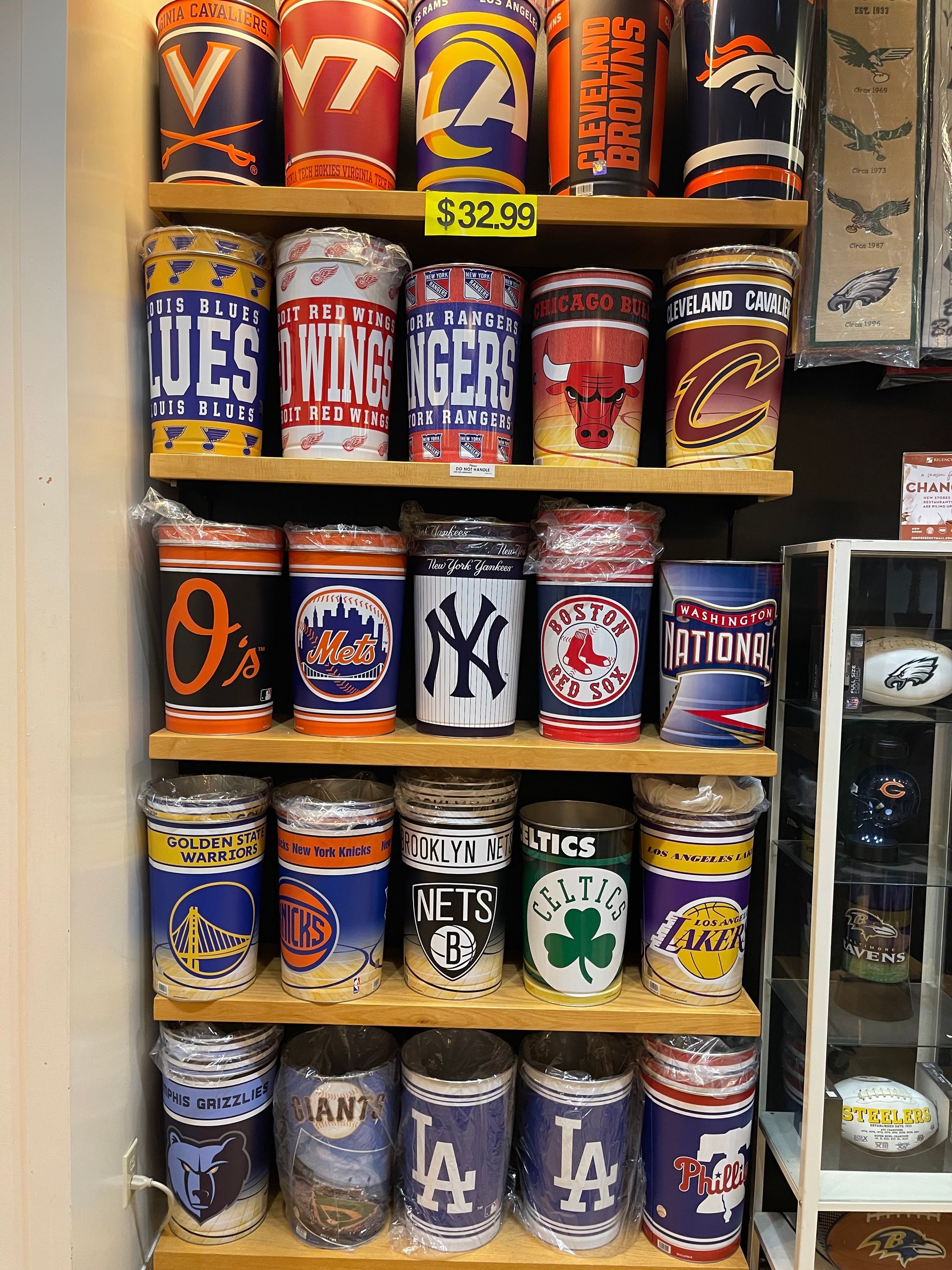 A retail display shelf stocked with various sports team tumblers featuring logos from the NBA, MLB, NHL, and college teams.