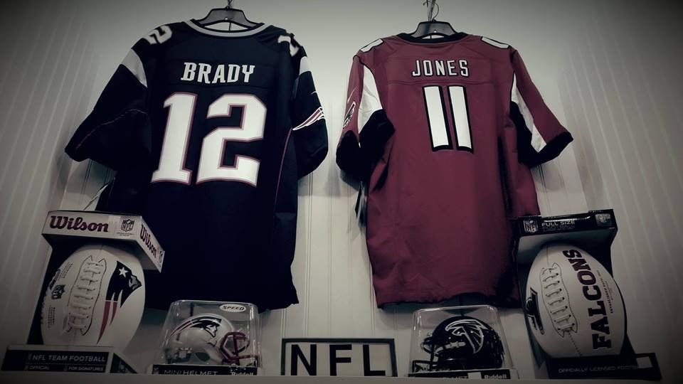 Two NFL jerseys, Brady 12 and Jones 11, displayed above two footballs and two miniature helmets on a shelf.