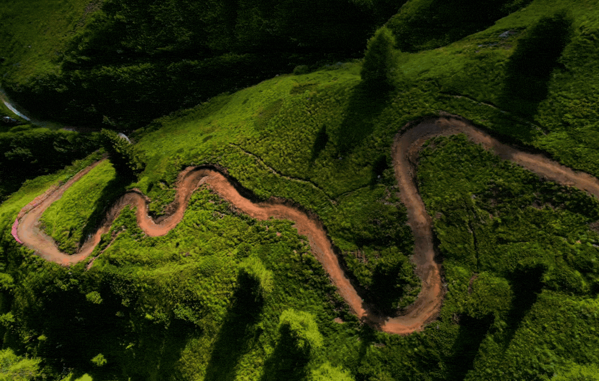 A winding dirt road cuts through lush green hills and trees from an aerial view with a Pathfinder logo overlay