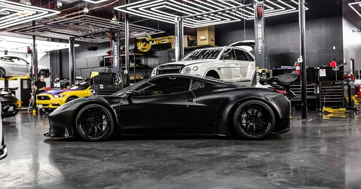 Black sports car in modern automotive workshop with tools, lift equipment, and white vehicle on hoist overhead.