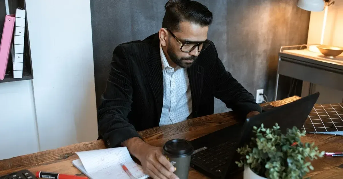 Professional man in black blazer reviewing car detailing keywords on laptop at wooden desk workspace.
