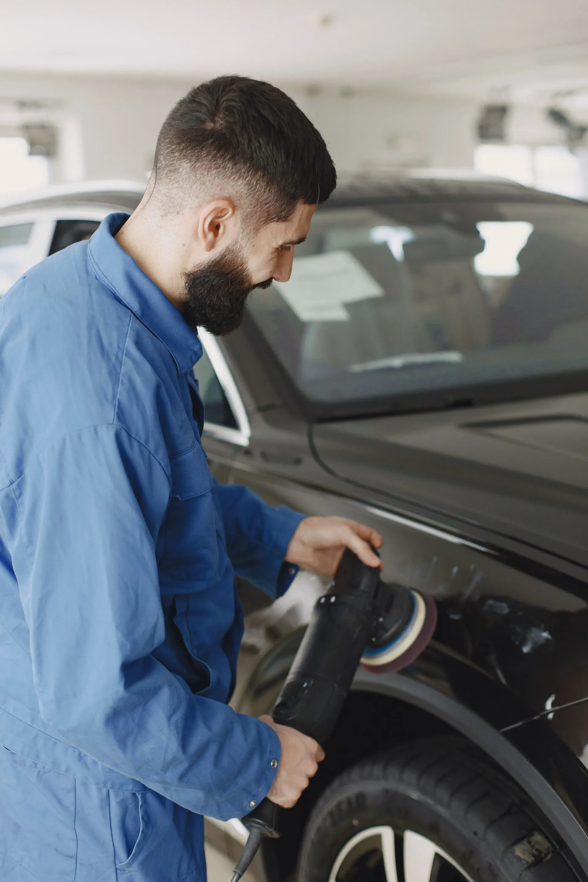 A man in a blue jumpsuit polishes a black car's hood using a power buffer in a well-lit garage, focusing intently on his task.