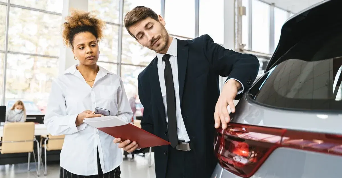 Car salesman in suit showing vehicle features to female customer with clipboard in a dealership.