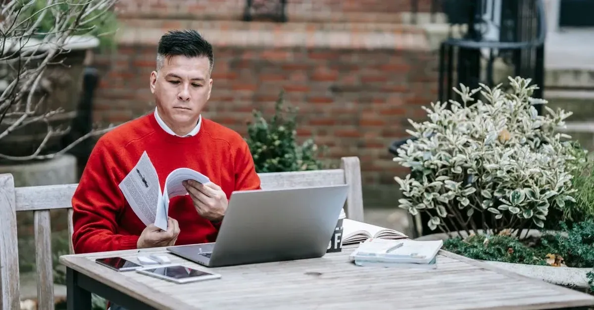 Man in red shirt working on laptop at outdoor table with plants and brick wall background.