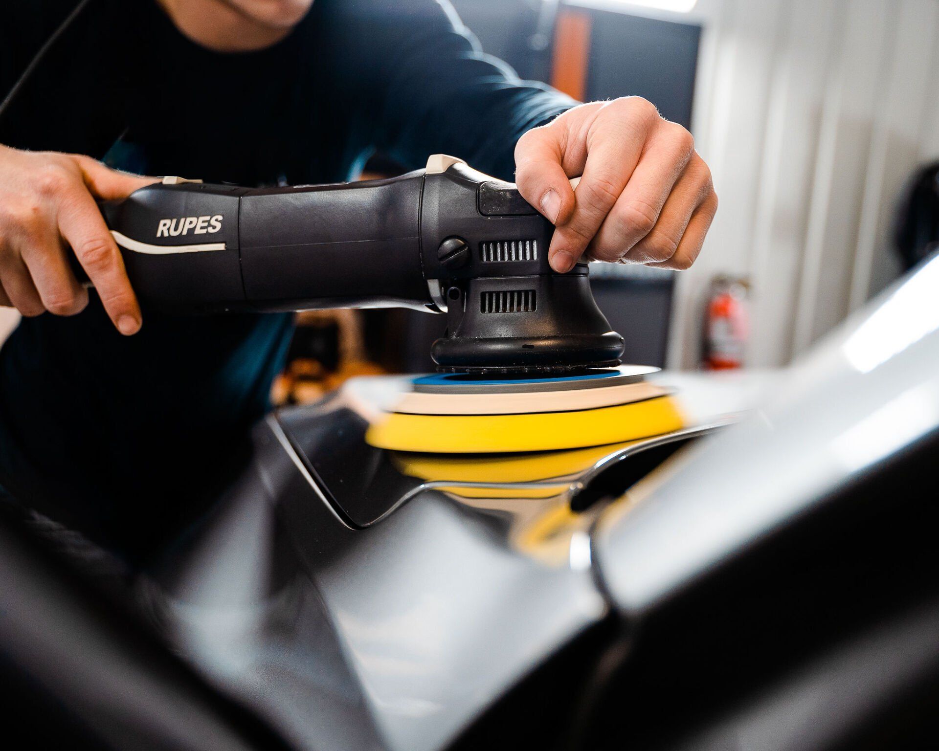 Person polishing a car with a black Rupes polisher and yellow/blue pad.