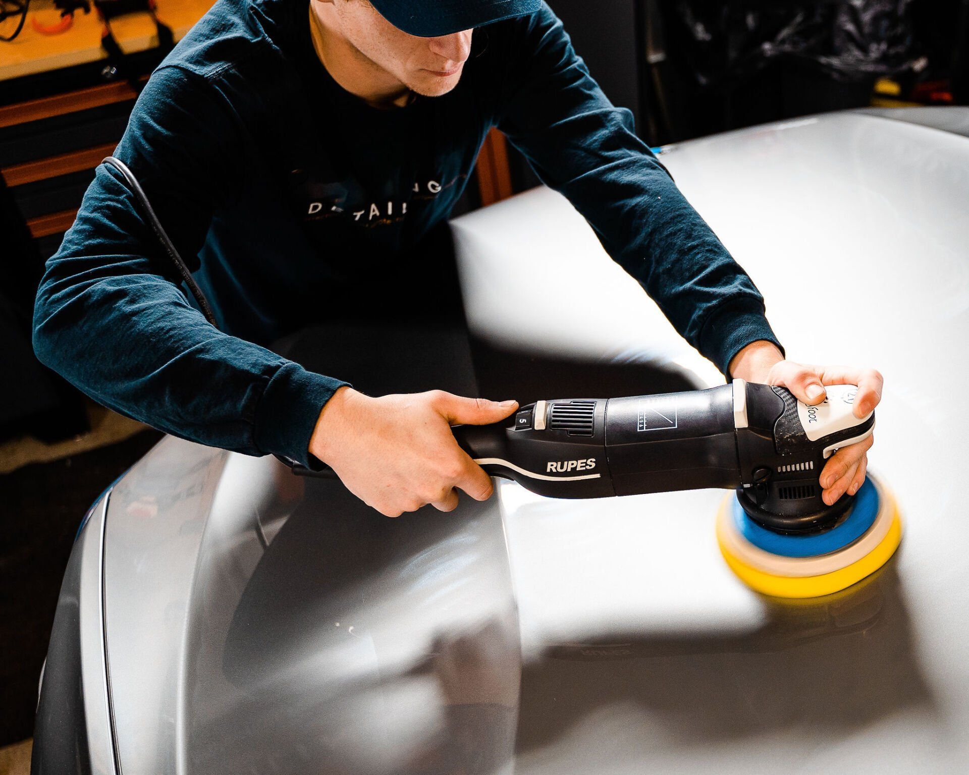 Person polishing a car's gray surface with an electric buffer, in a workshop.