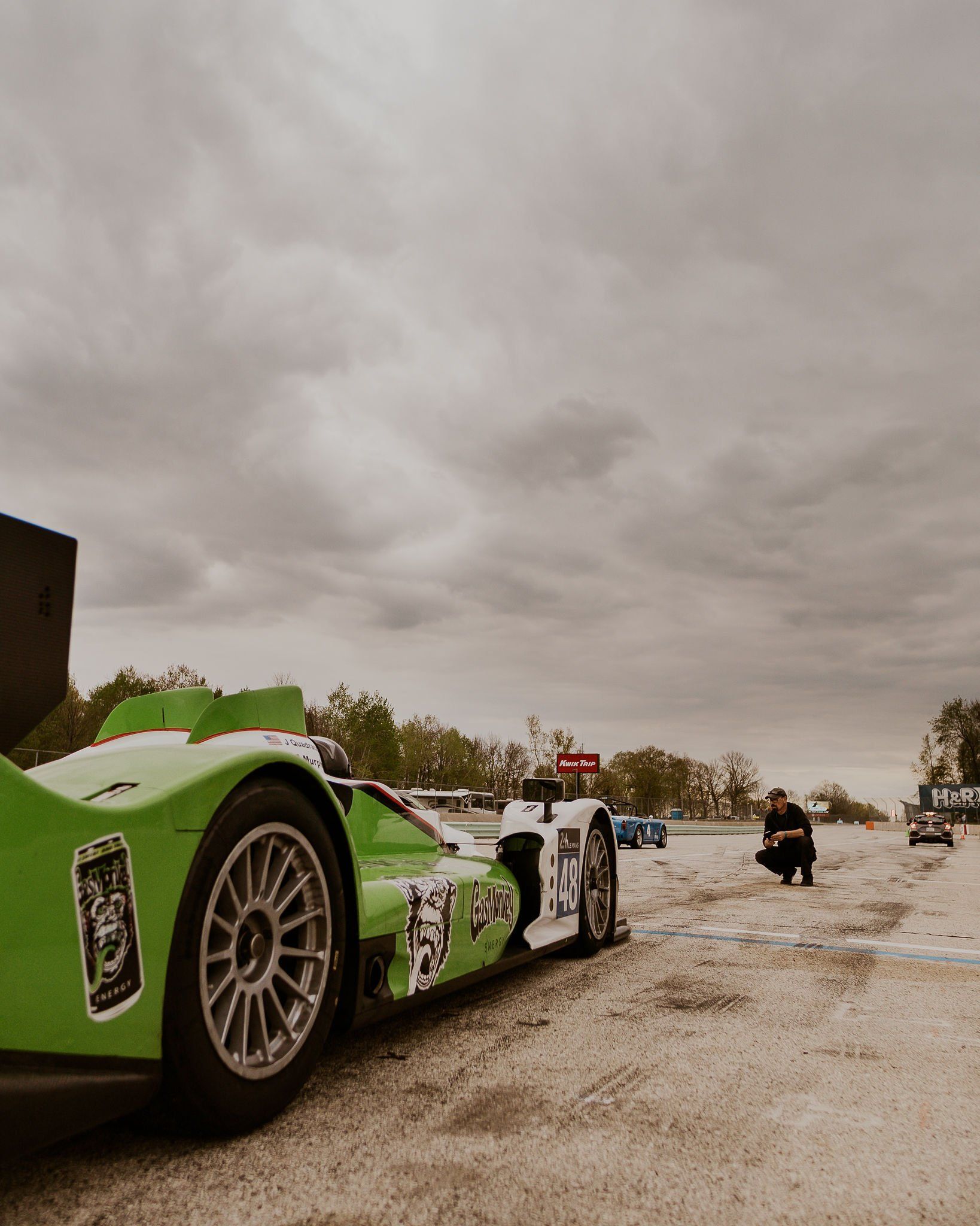 Green race car on a track with a mechanic squatting nearby under a cloudy sky.
