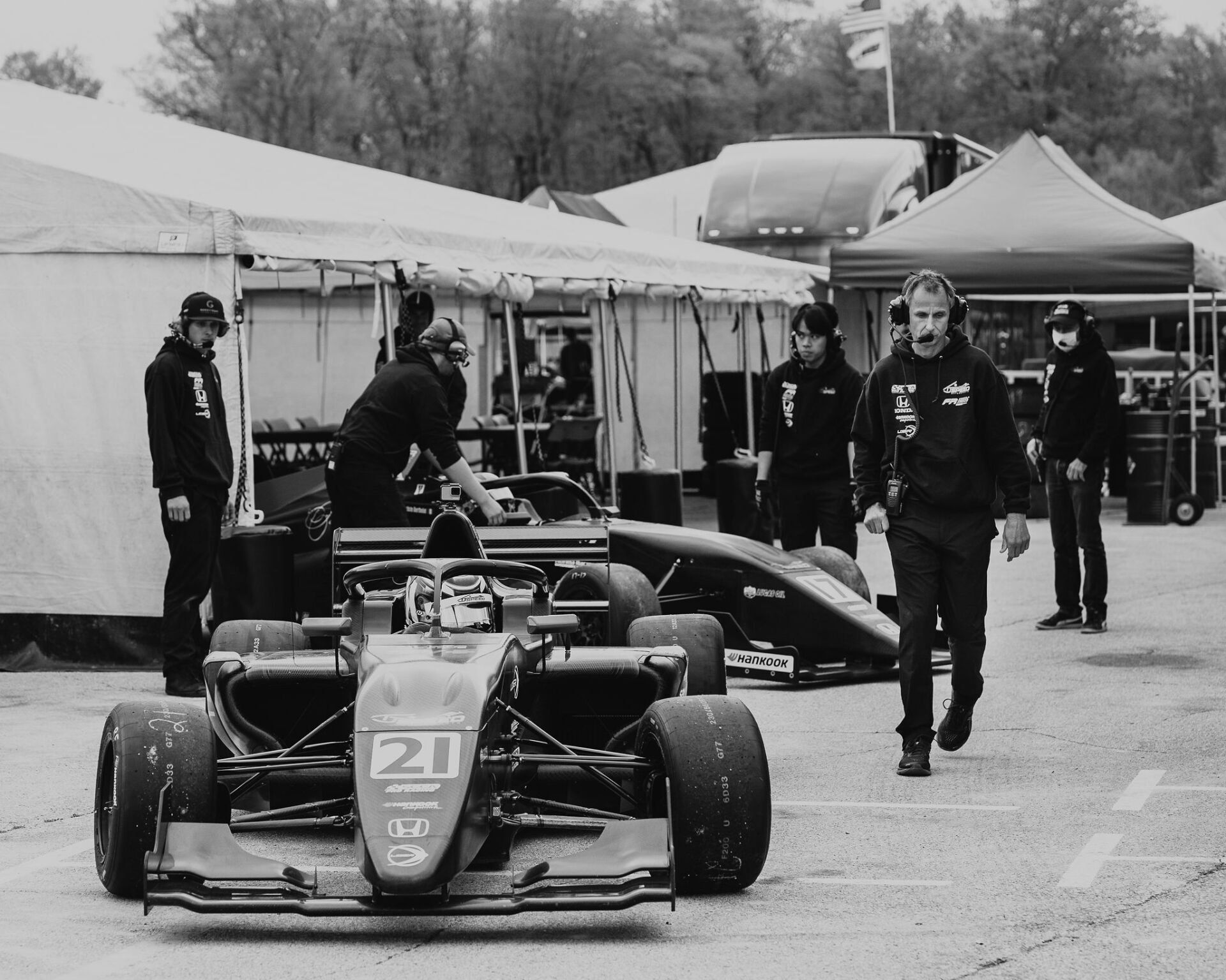 Race car in pit lane, mechanics working, other team members walking. Black and white.