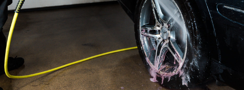 A person uses a yellow hose to wash a car's silver alloy wheel with pink foam.