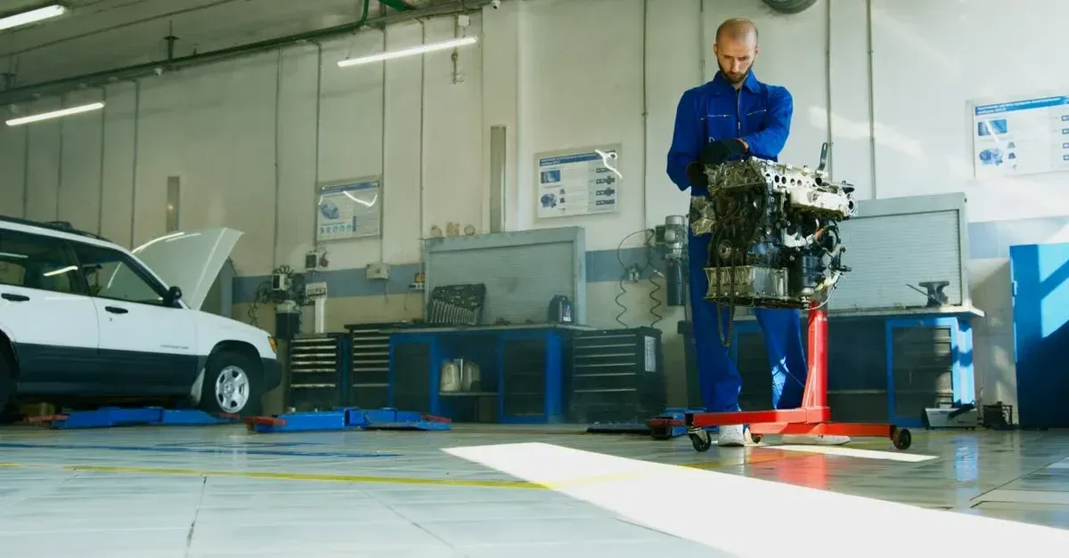 Mechanic in blue uniform holding car engine in professional auto repair shop with tools and white vehicle.