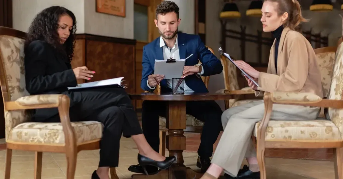 Three professionals discuss multi location marketing strategy using tablet and documents in modern office setting.