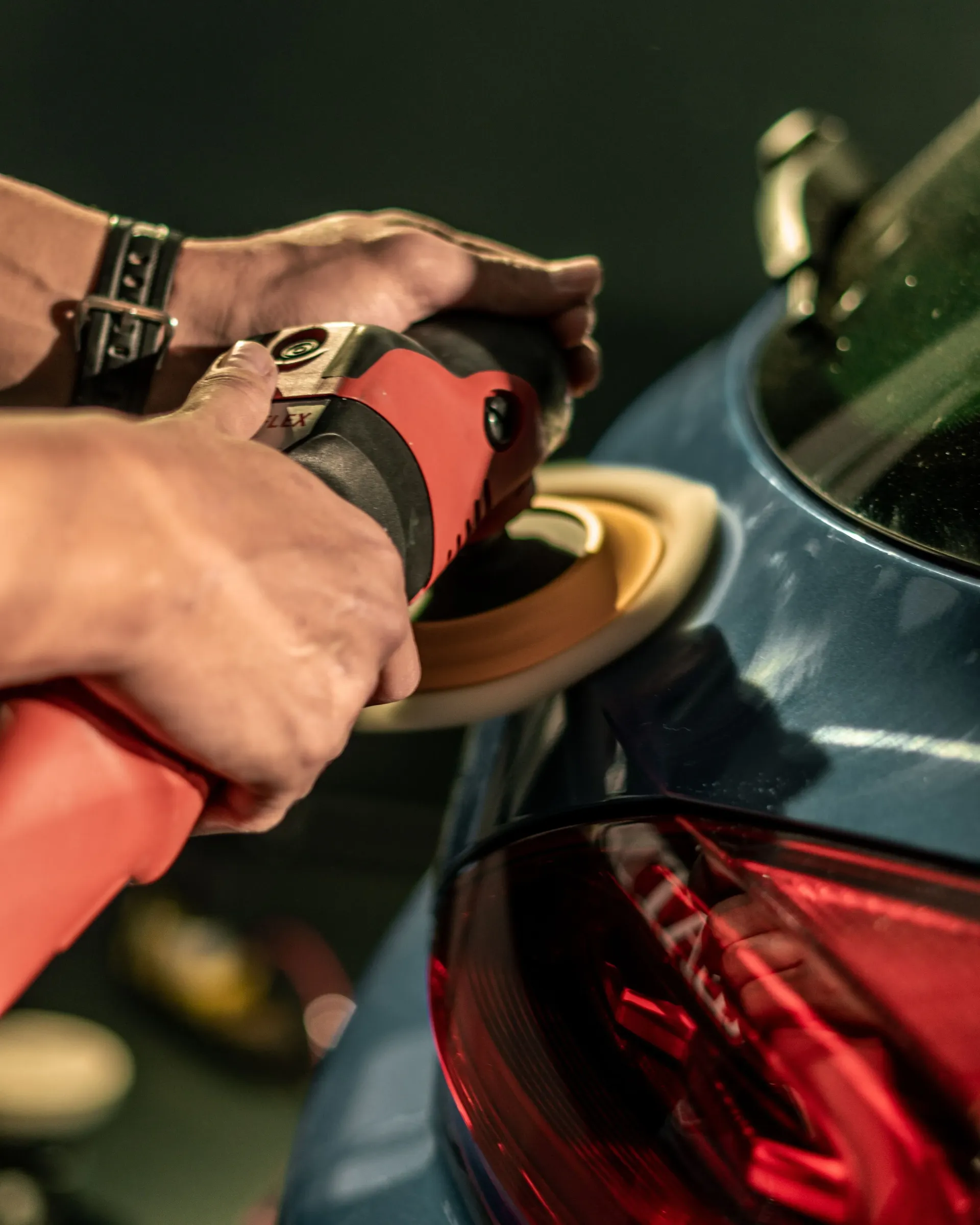 A person in a baseball cap works intently on a car with a tool in hand, the scene lit by focused lights, conveying concentration in a dim garage setting.