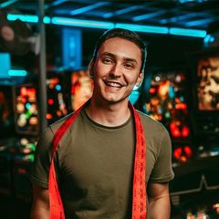 A man wearing a red scarf around his neck is smiling in front of a pinball machine.
