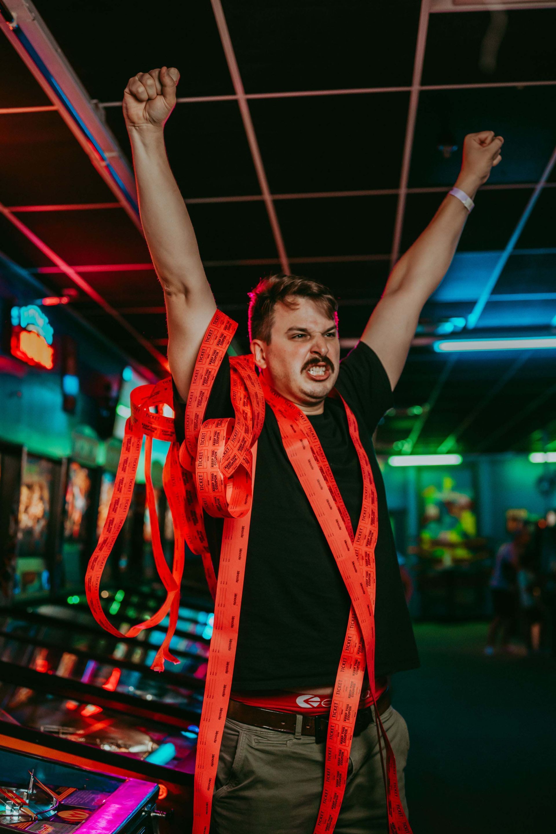 A man with a mustache is standing in an arcade with his arms in the air.