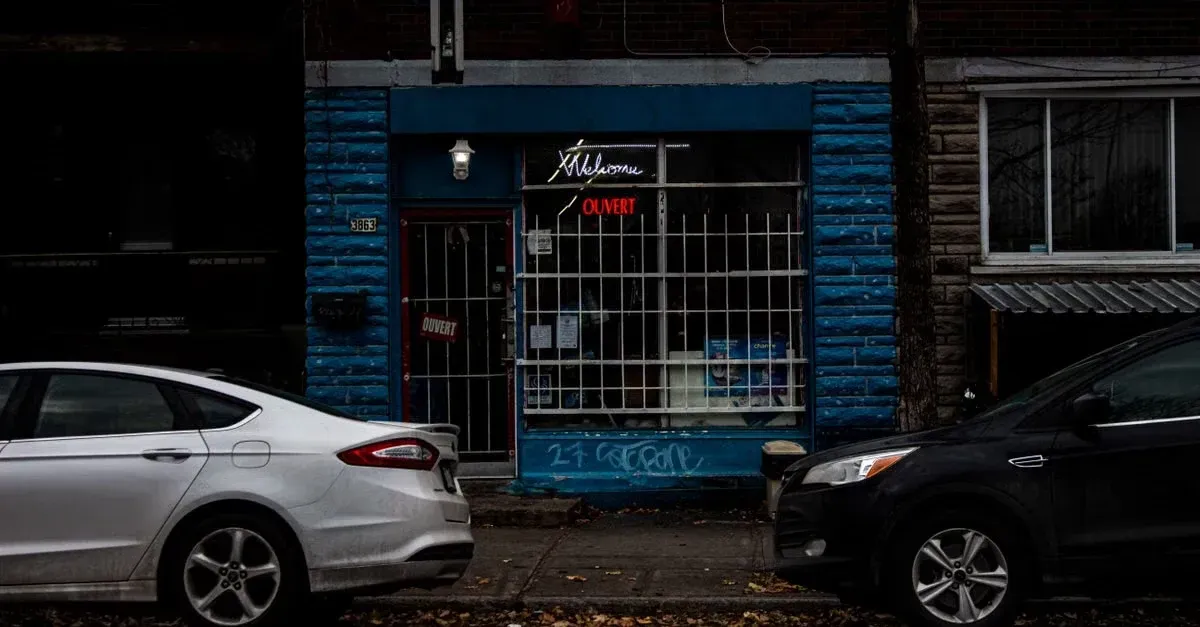 Blue storefront with metal grill gate between parked silver and black cars on street.