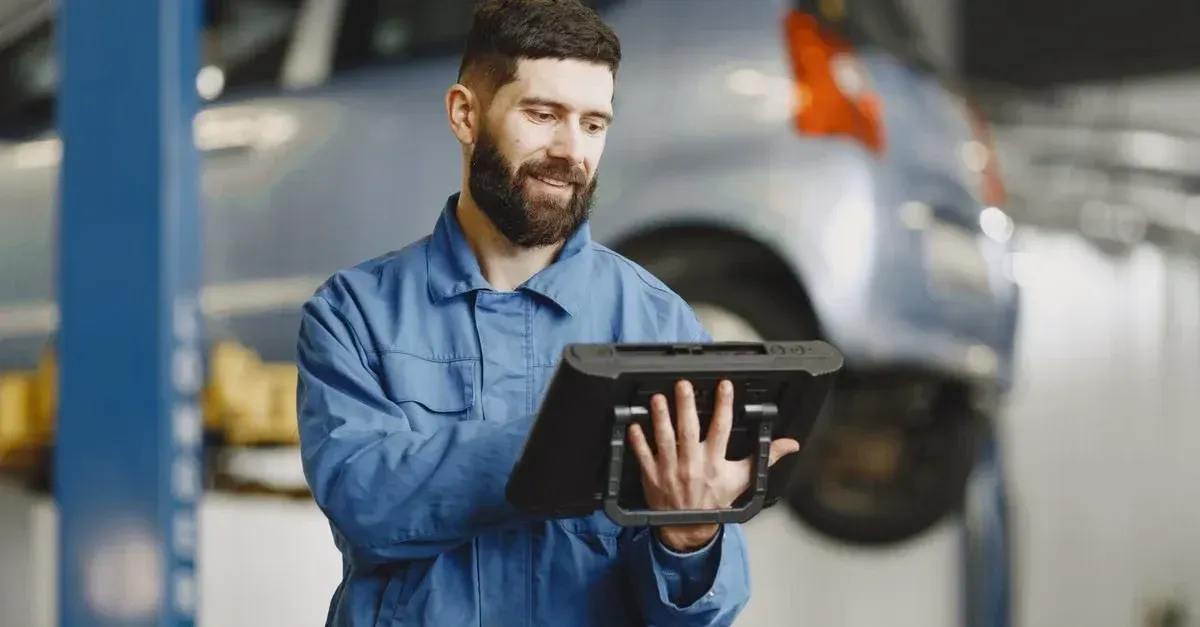 Mechanic in blue uniform holding tablet in auto repair shop with vehicles in background.