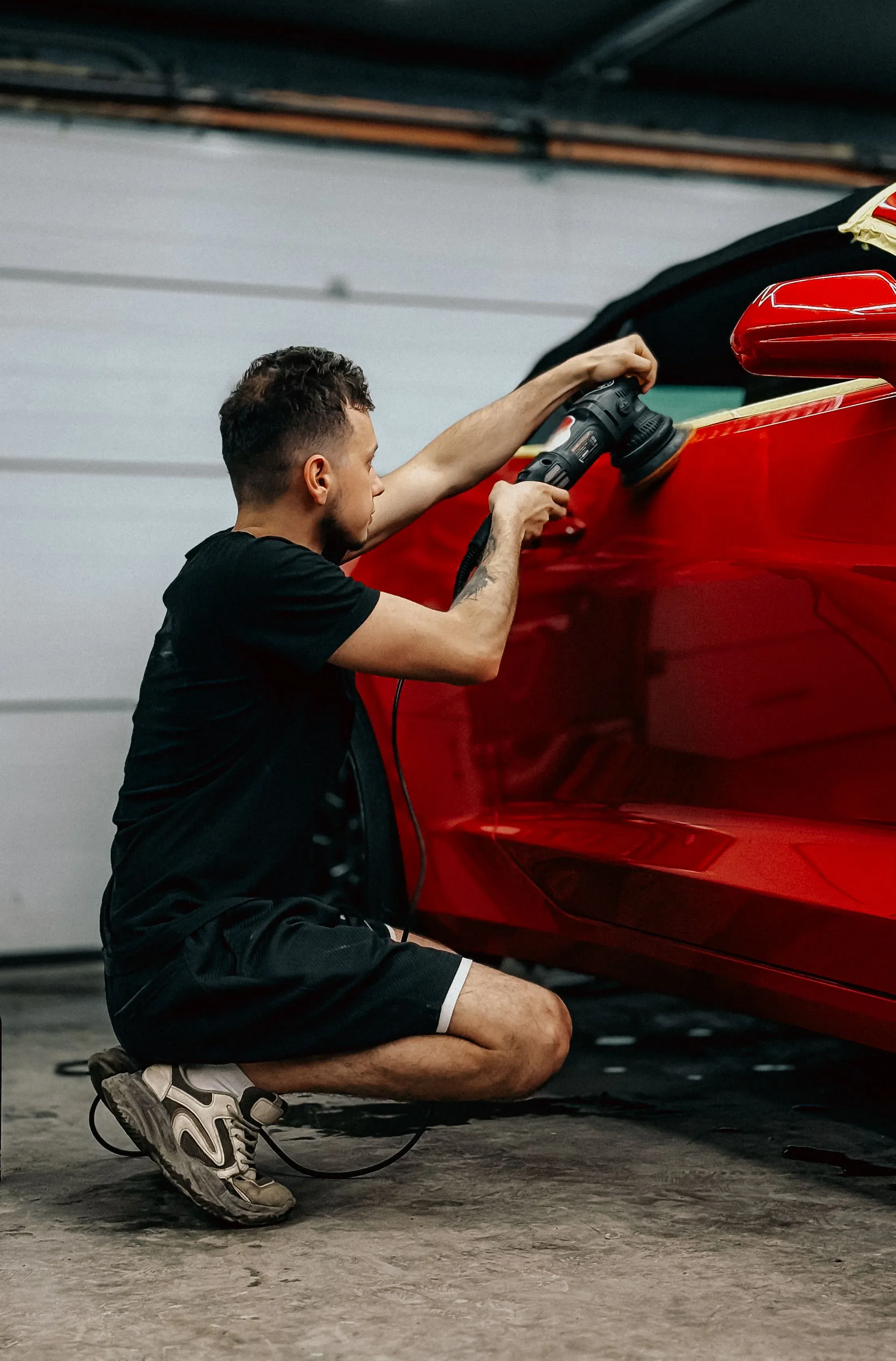 A technician in a black t-shirt and shorts uses an electric polisher to buff the glossy red side panel of a car inside a professional garage shop.