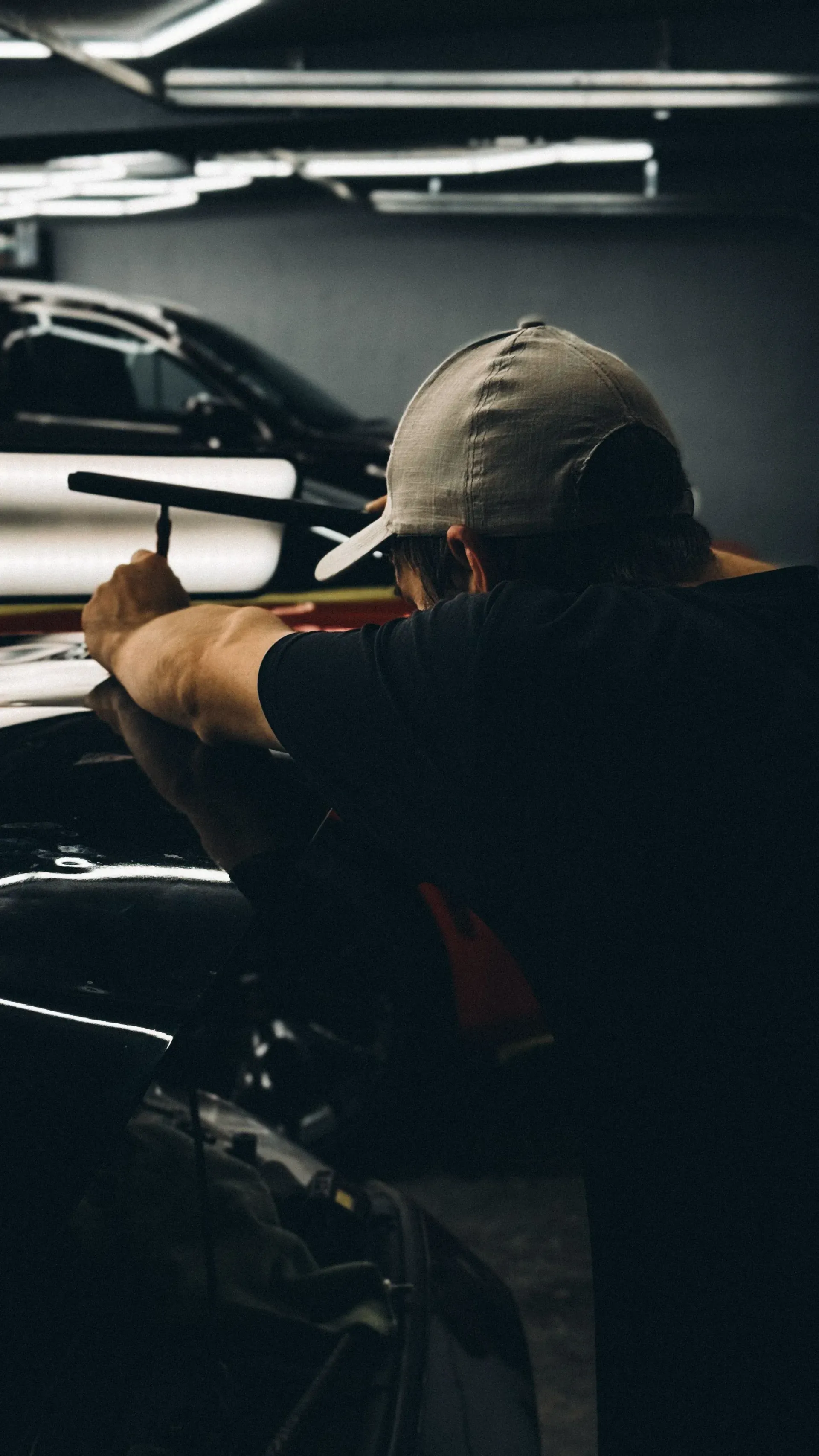 A person in a baseball cap works intently on a car with a tool in hand, the scene lit by focused lights, conveying concentration in a dim garage setting.