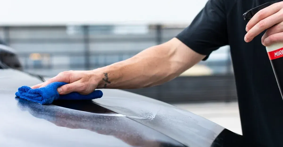 Worker applying car detailing business protective coating with blue microfiber cloth on vehicle surface.