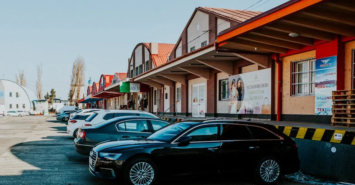 A row of parked cars lines storefronts beneath red-orange awnings in a quiet commercial plaza, with clear skies and a calm, everyday atmosphere.