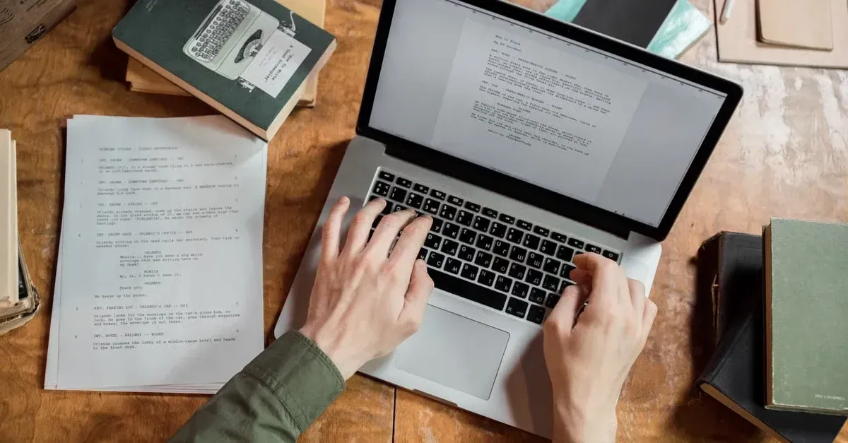 Hands typing on laptop at wooden desk surrounded by books and printed documents while writing website content.