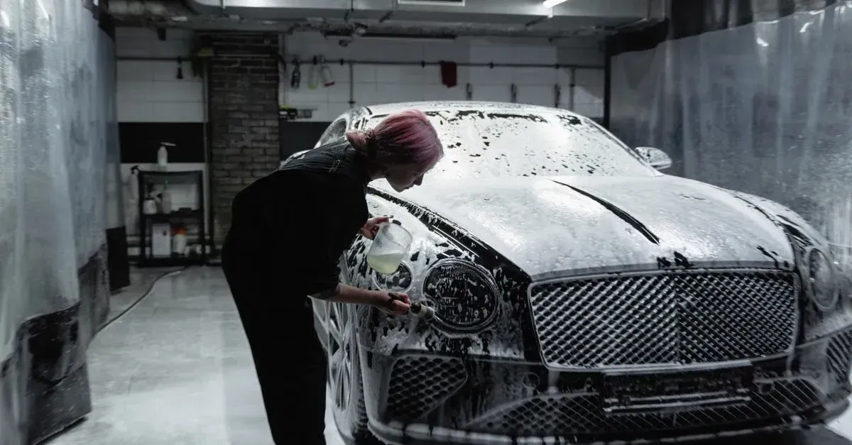 Professional detailing technician washing luxury car hood with foam in industrial garage setting.