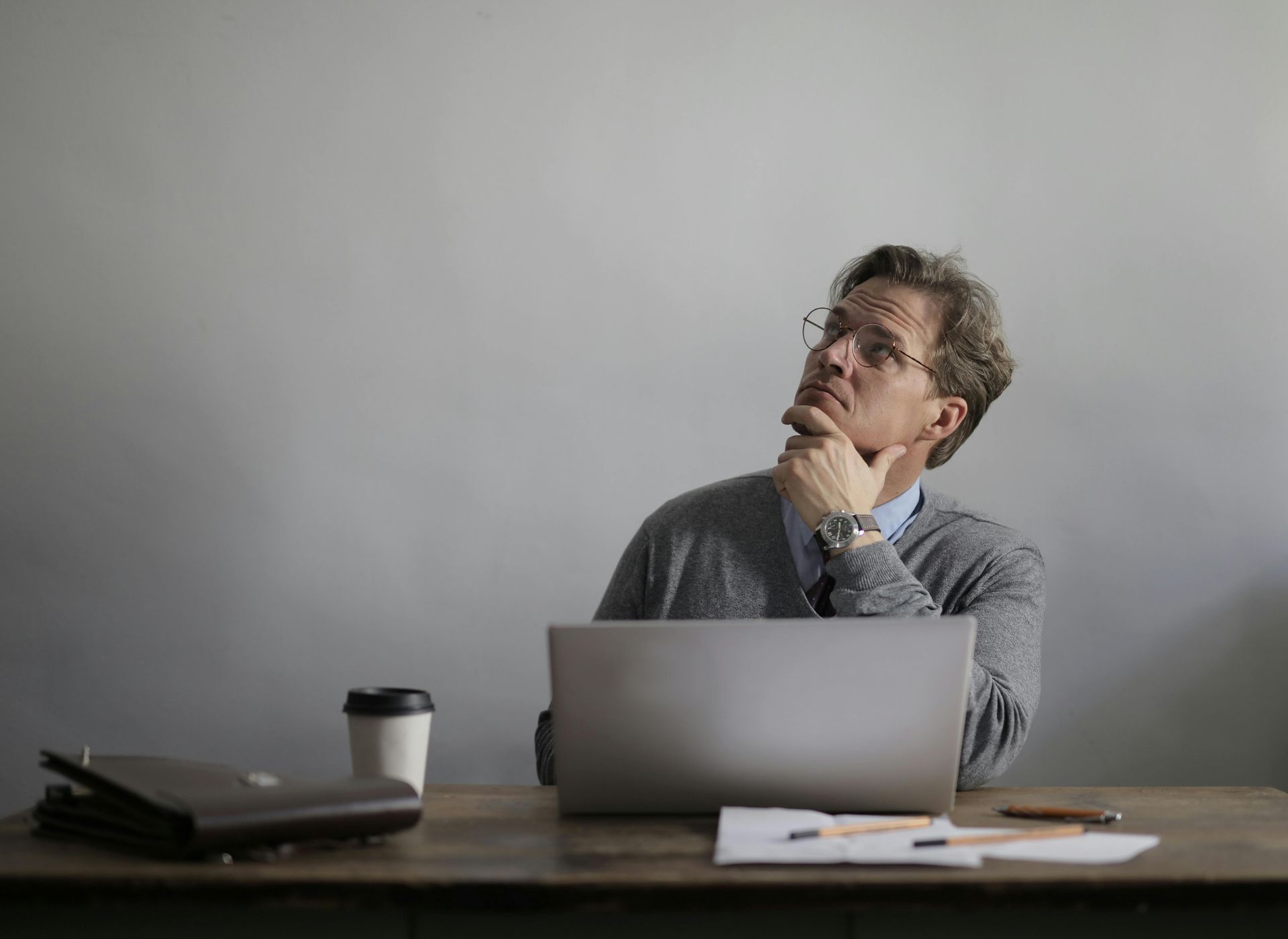 Person thinking at a desk with a laptop and coffee cup, looking up in a minimalist office.