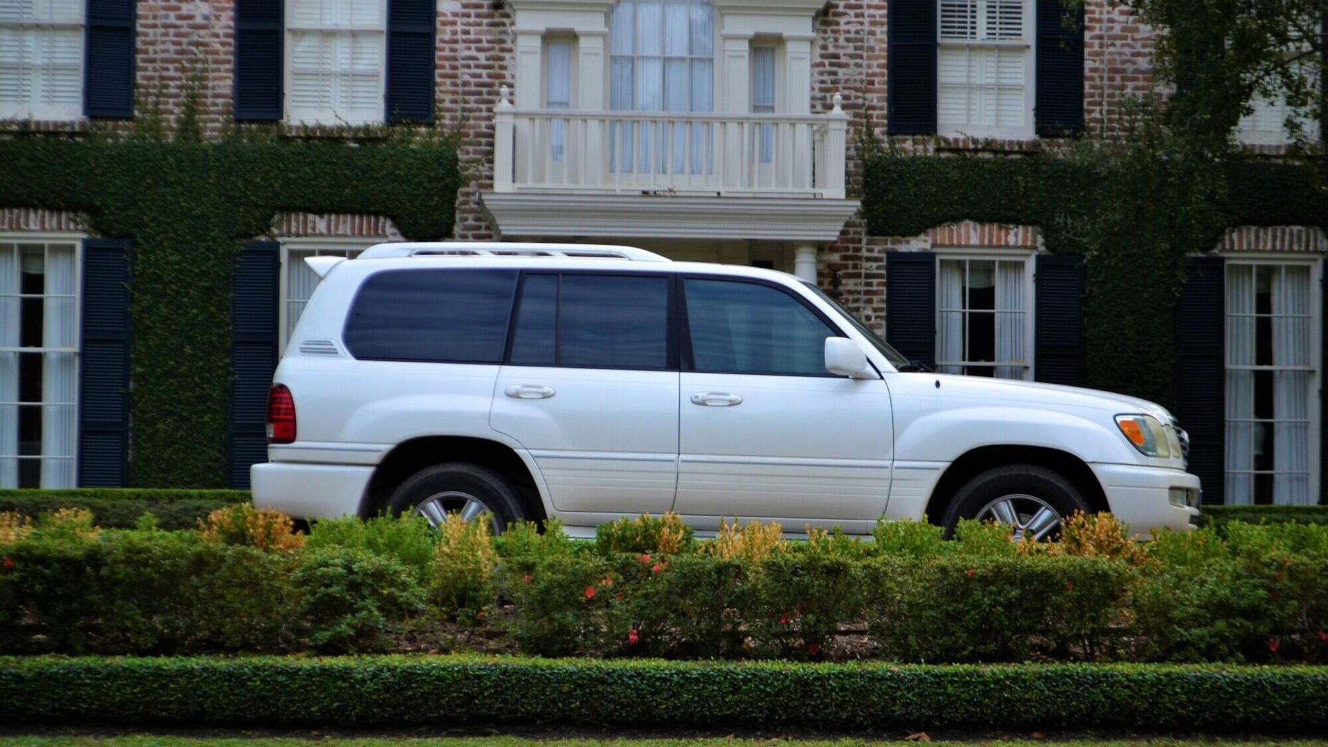White SUV parked in front of a brick house with dark shutters, surrounded by manicured hedges.