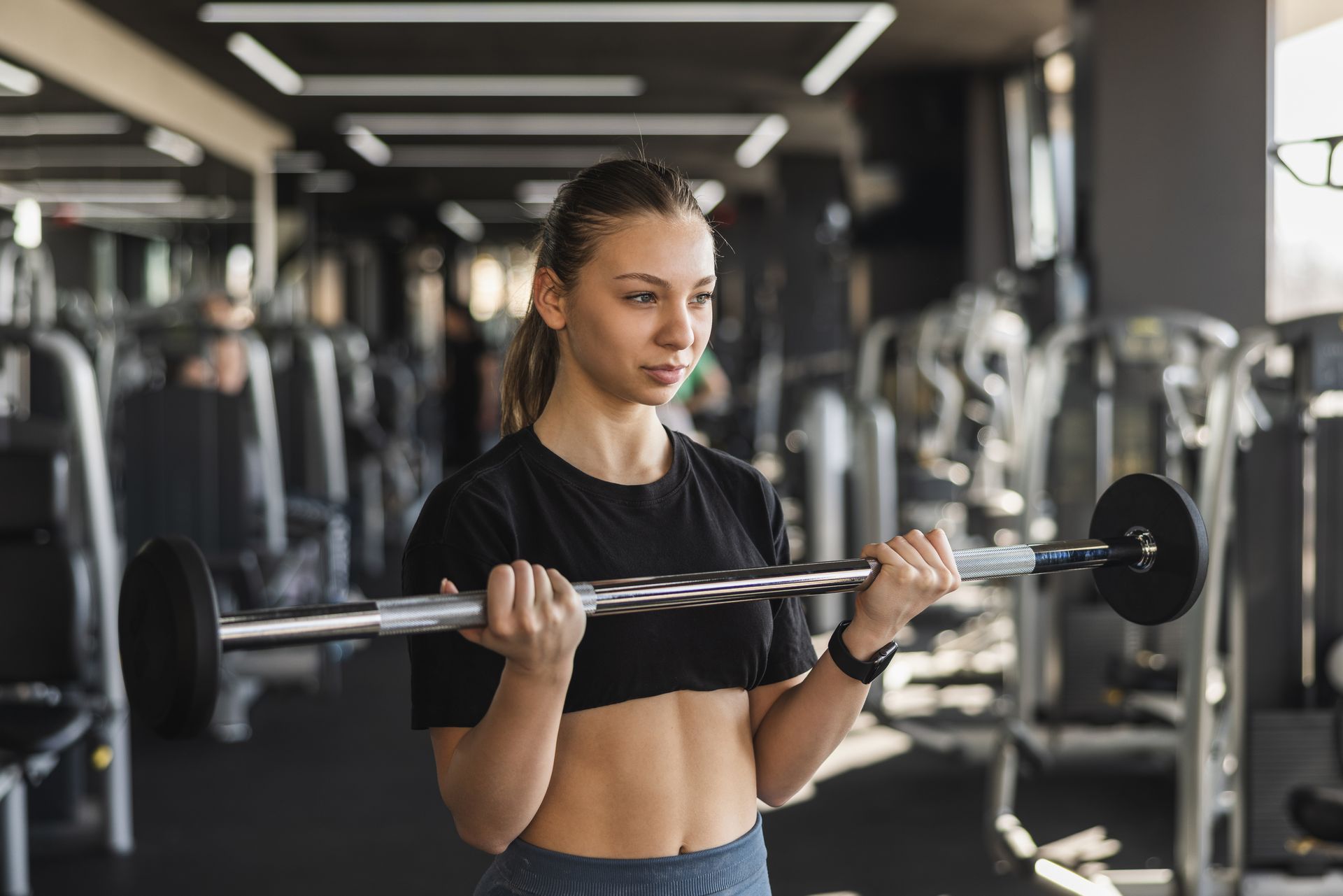 Woman lifting a barbell in a gym, wearing a black crop top and focused expression.