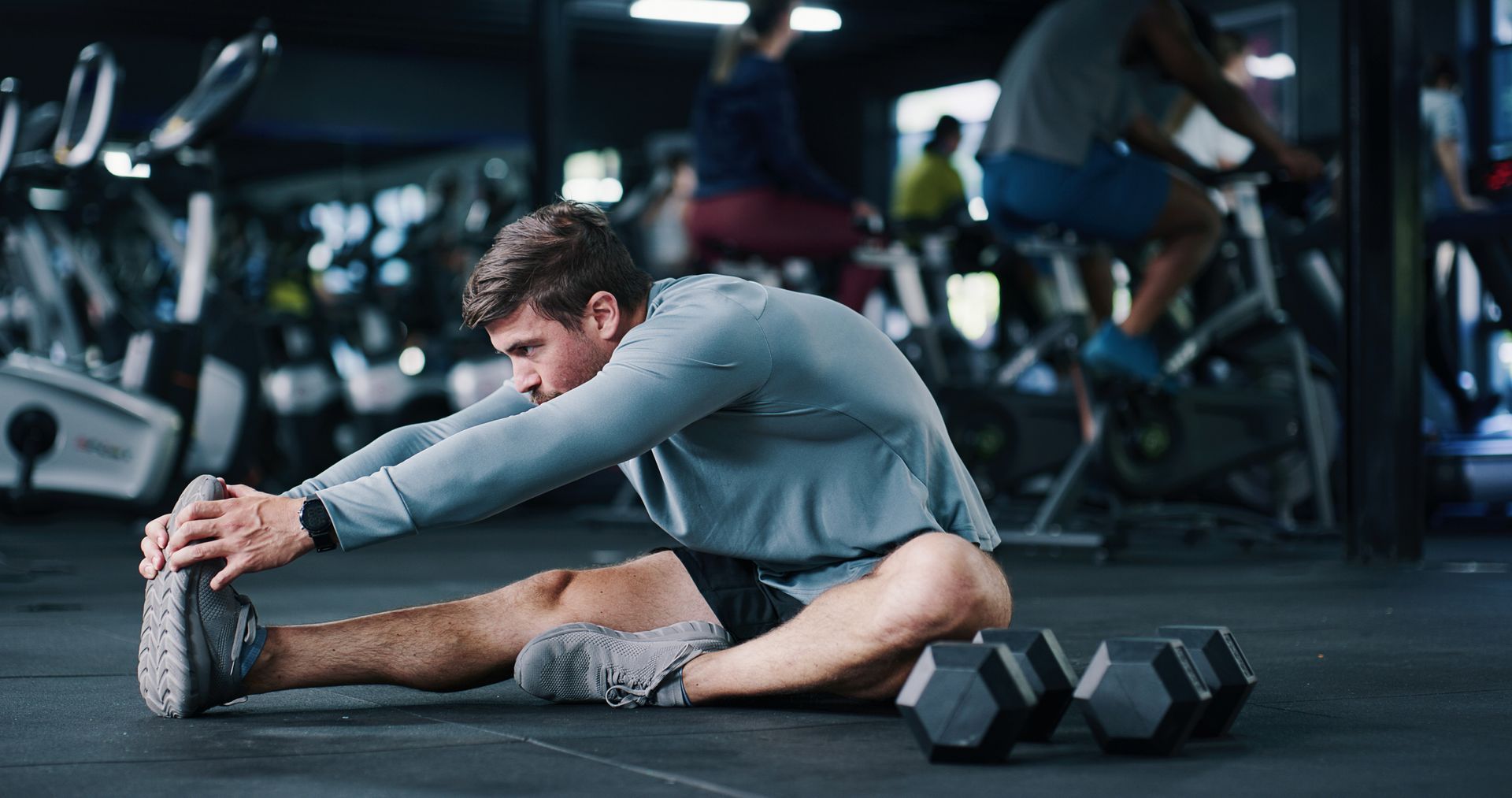 Man stretching on gym floor beside dumbbells, reaching toward his foot
