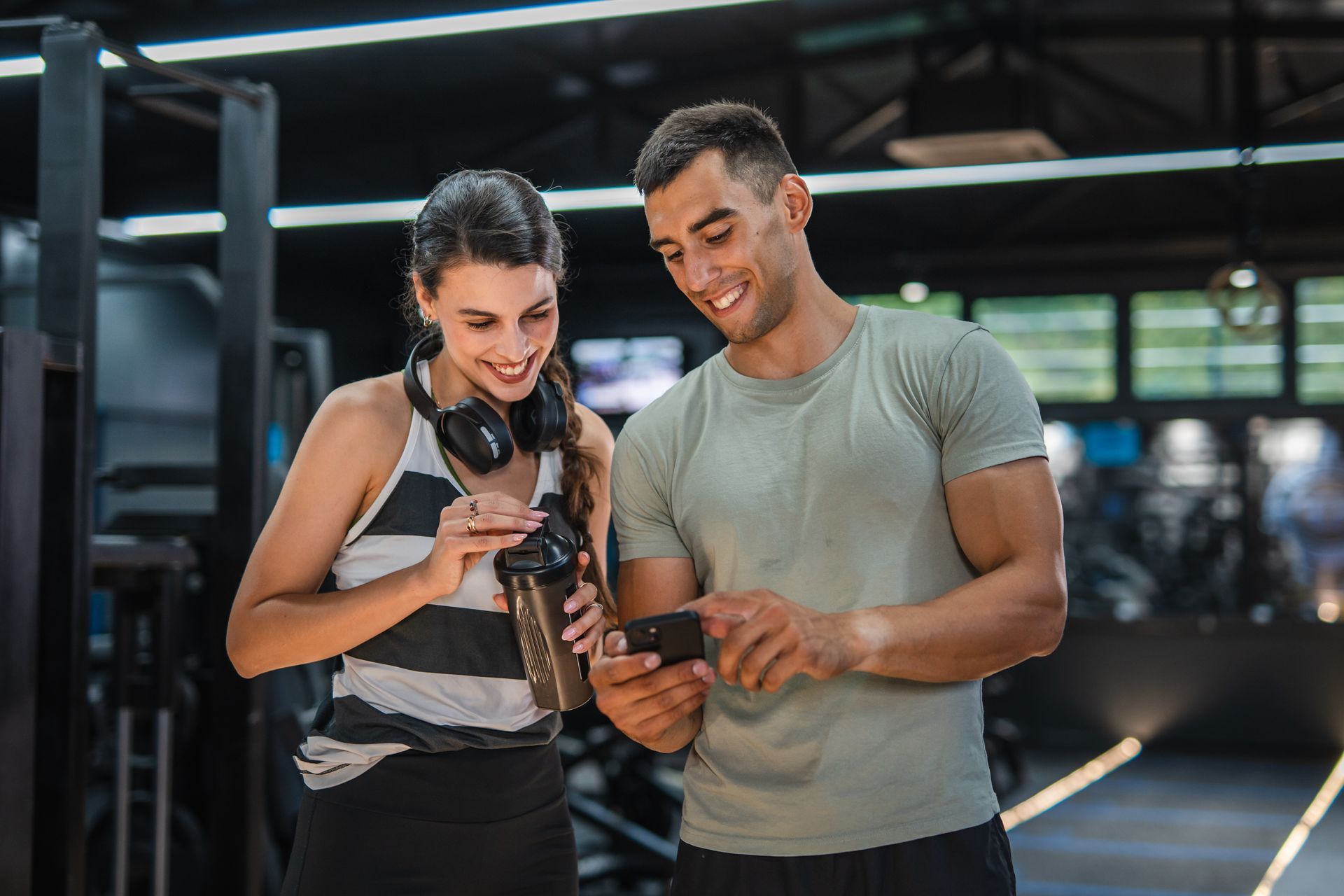 Two people smiling and looking at a camera in a gym, with one holding a phone and the other pointing at it
