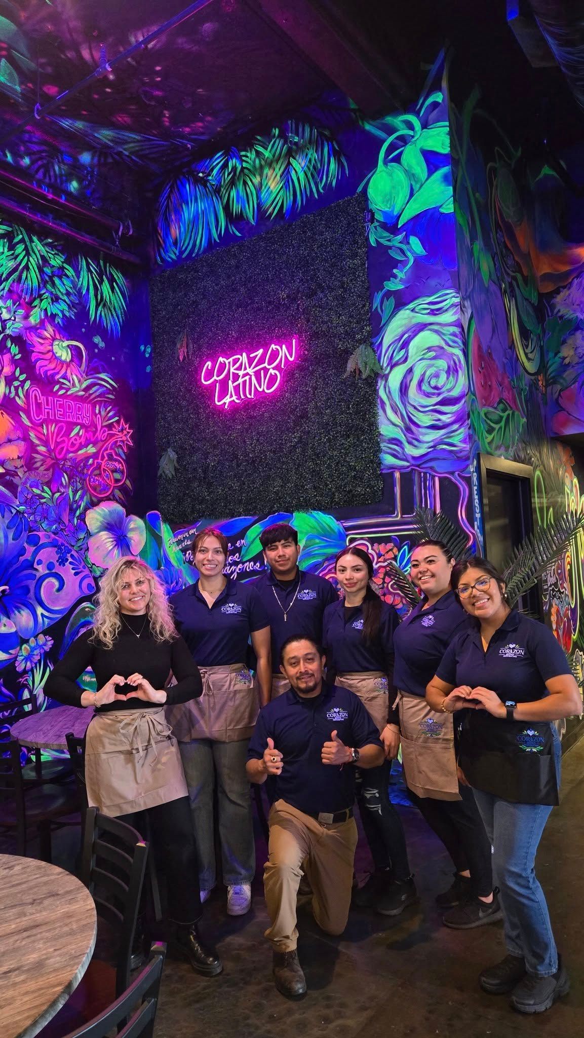 Group of restaurant staff posing, hands in heart shapes. Fluorescent art in background.