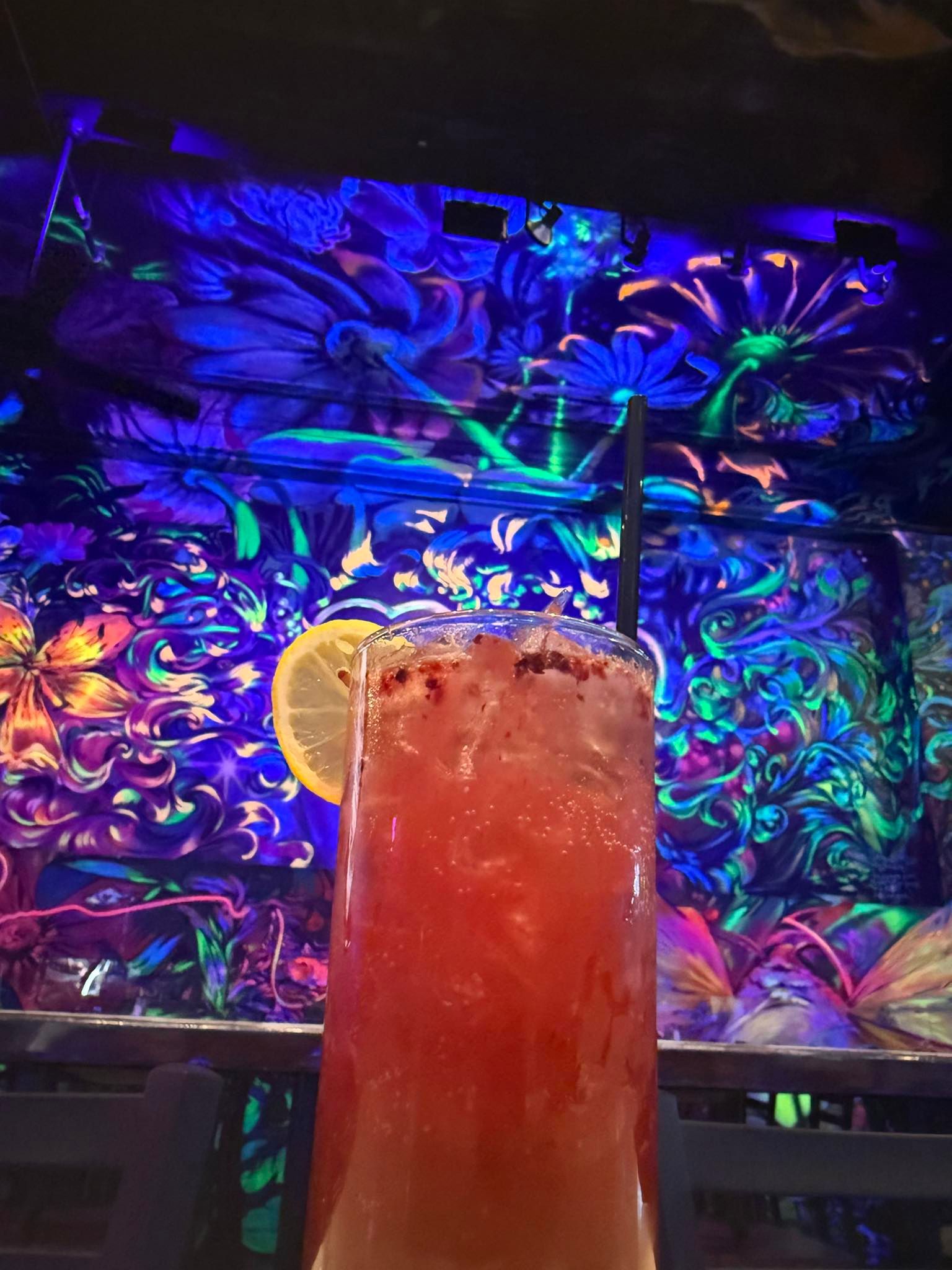 Tall cocktail with lemon slice and straw in front of a neon floral mural.