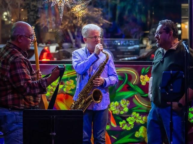 Three musicians performing. A saxophone player sings, a guitarist plays, and another man looks on. Colorful background.
