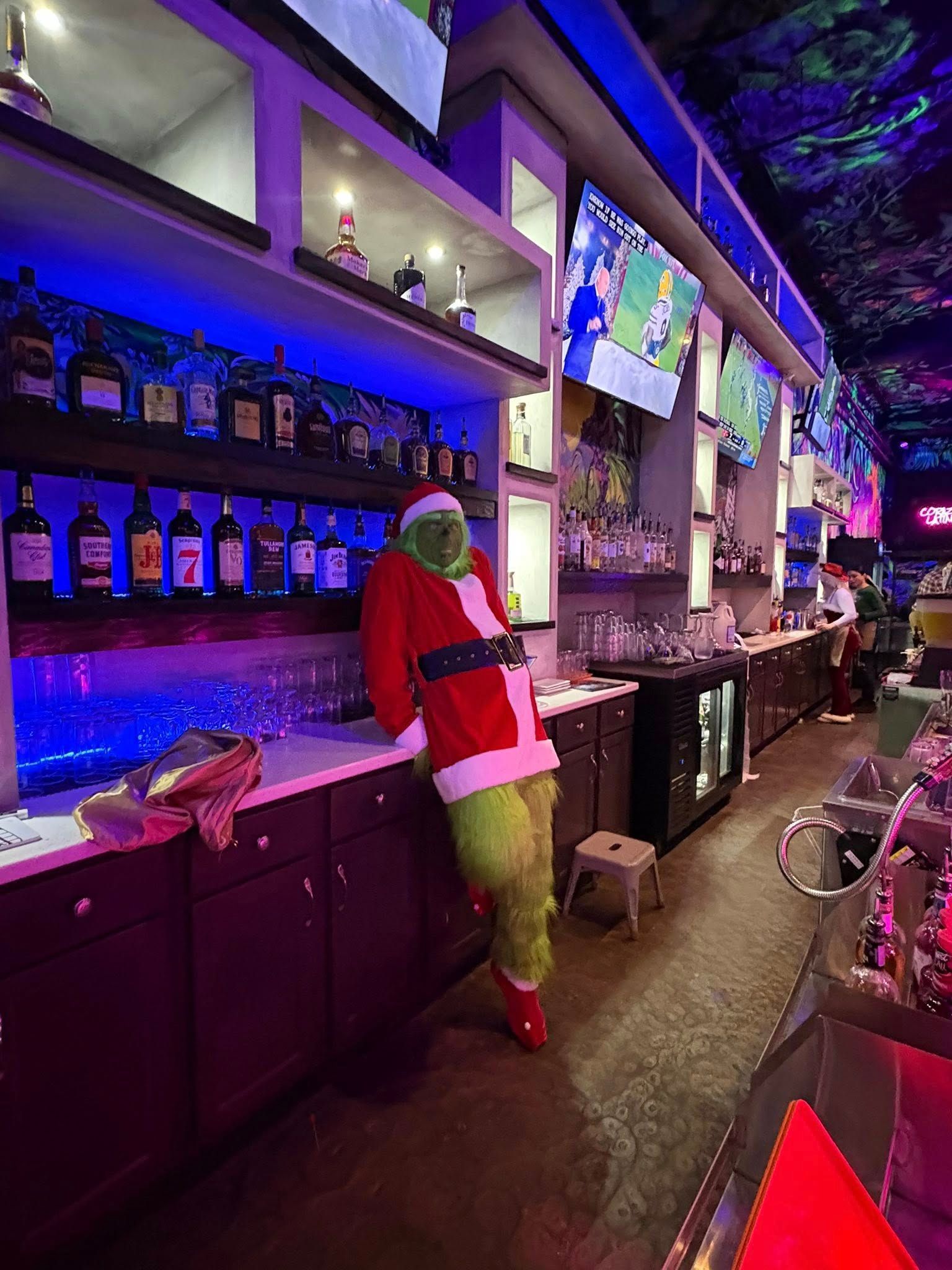 The Grinch in Santa suit leans against a bar, lit with blue lights, with liquor bottles and TVs visible.