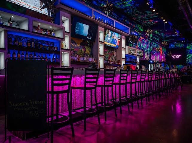 Bar with pink and blue lights, chairs, and shelves of liquor. TVs above bar.