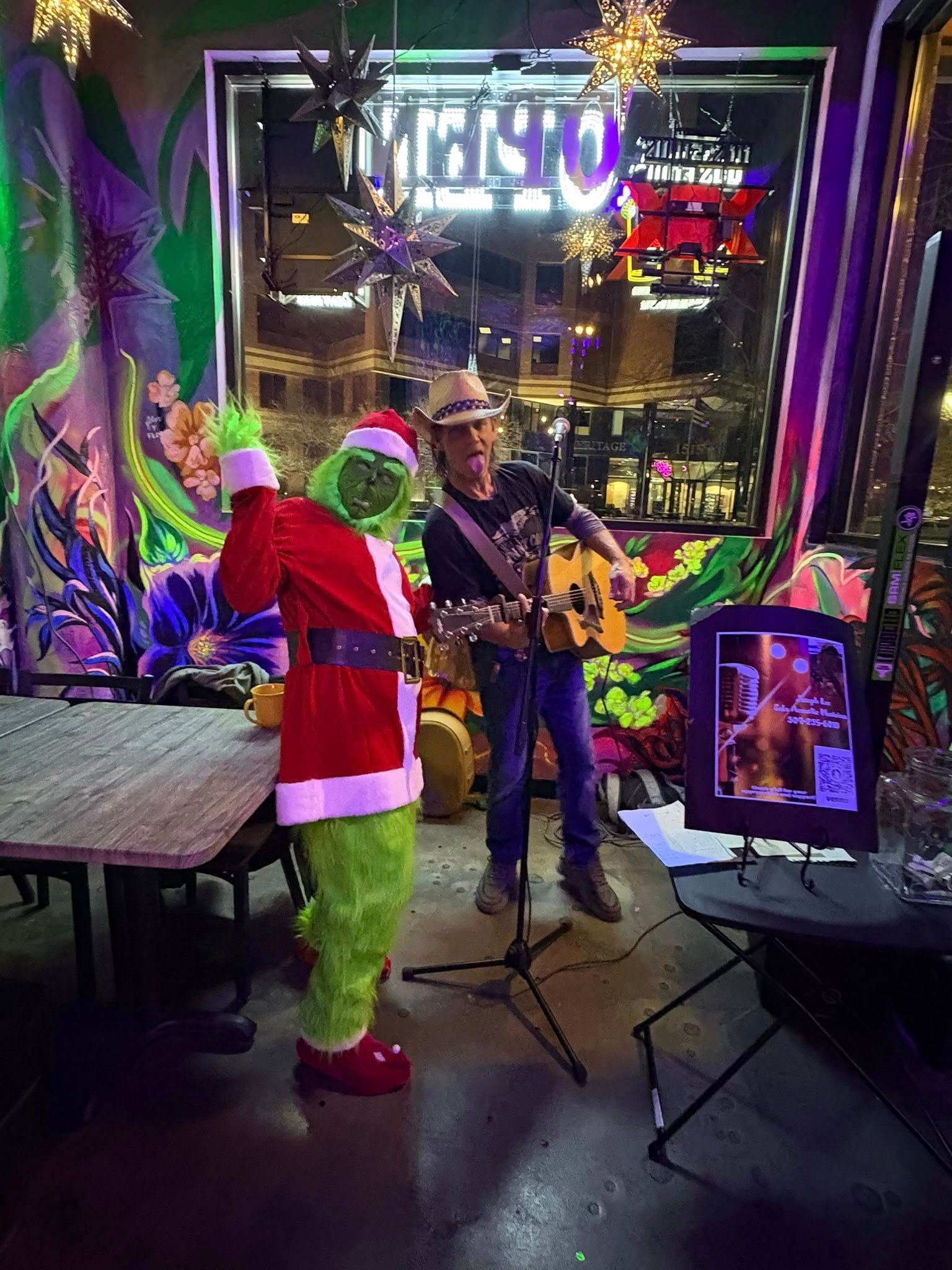 Grinch in Santa suit dances near a musician playing guitar in a dimly lit bar, with city lights visible outside.